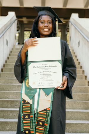 Graduate in black cap and gown holding diploma on outdoor staircase with kente cloth stole.