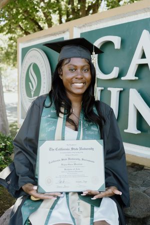 Smiling graduate in cap and gown holding diploma in front of California State University Sacramento sign.