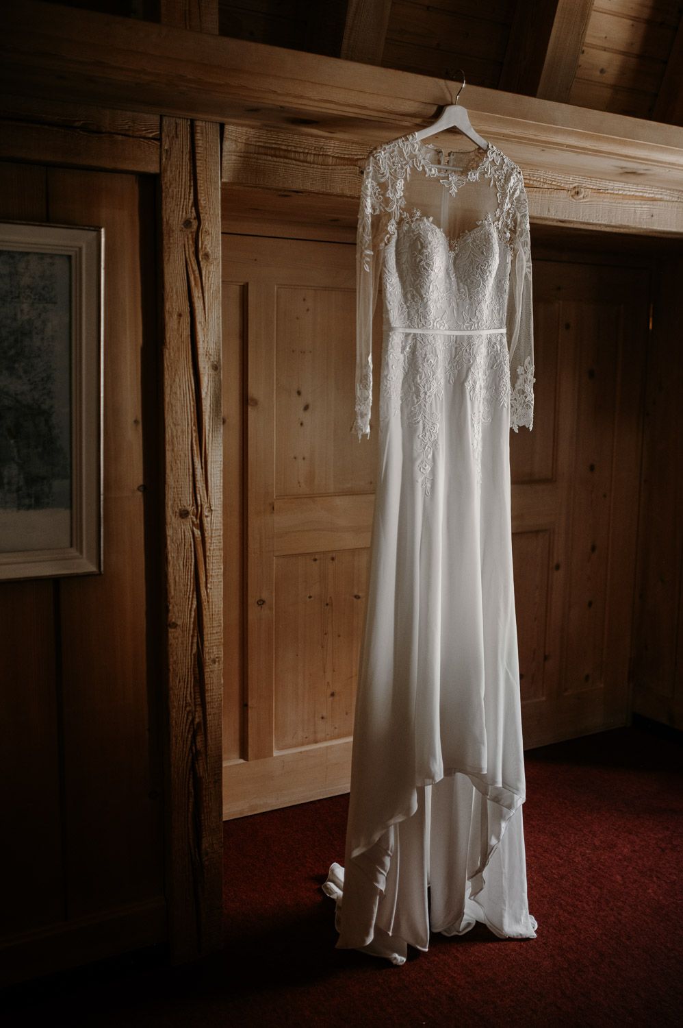 Elegant white lace wedding dress with long sleeves hanging on a wooden wall in a rustic cabin.