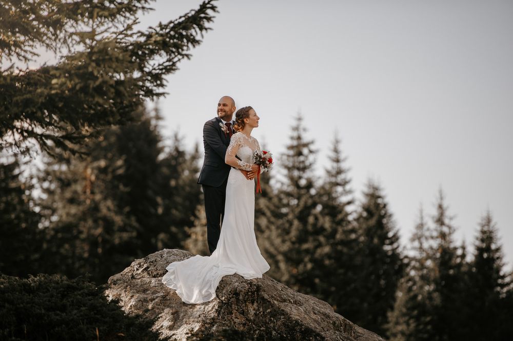 Bride and groom standing on a rocky mountain peak surrounded by evergreen trees during their outdoor wedding.