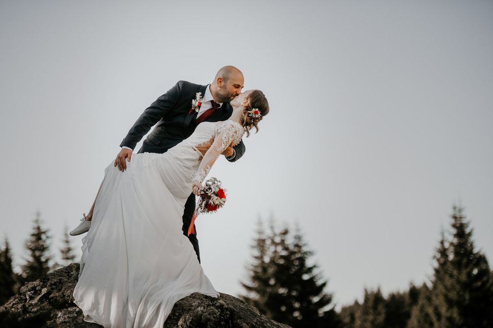 Groom dipping bride in white lace gown on rocky hilltop with evergreen trees behind them.