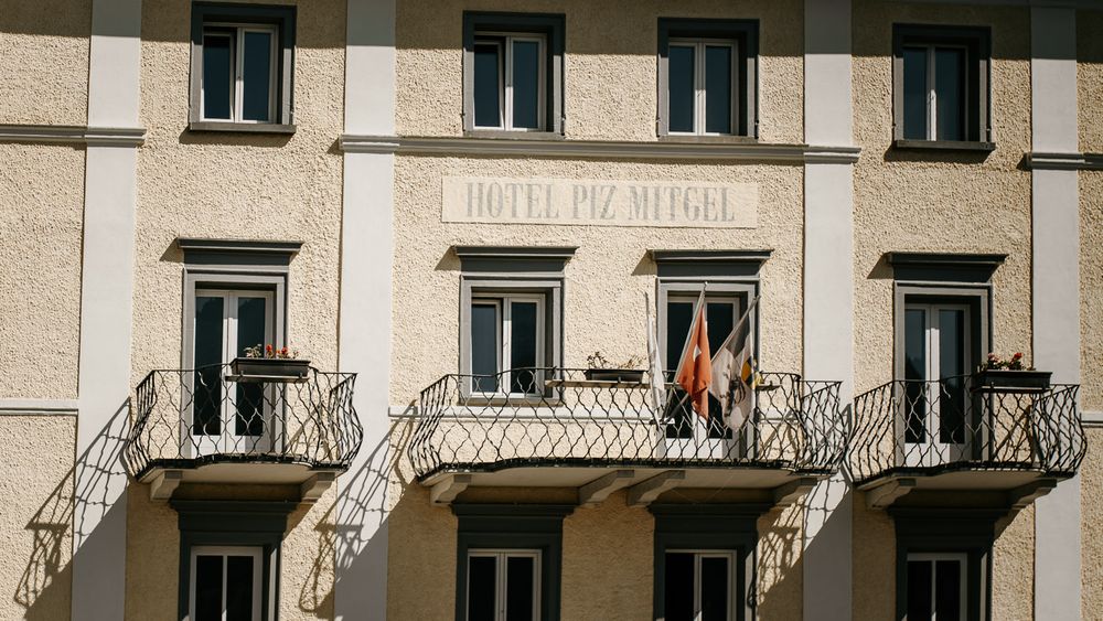Facade of Hotel Piz Mitgel with balconies and flags on a sunny day in the Alps.