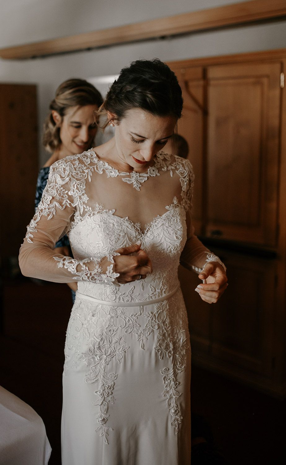 Bride in elegant lace long-sleeve wedding dress being helped by bridesmaid during bridal preparations.