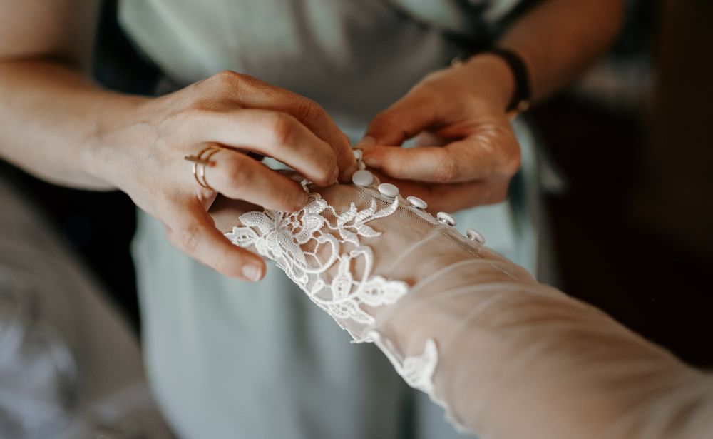 Close-up of bridesmaid fastening delicate lace buttons on bride's wedding dress sleeve.