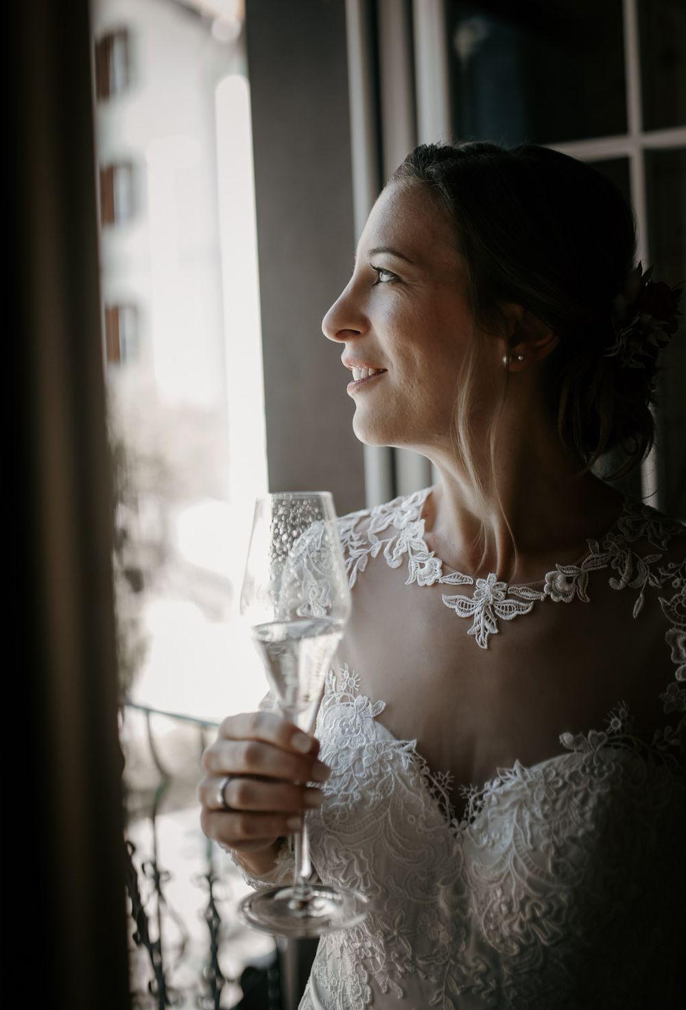 Smiling bride in lace wedding dress holding a champagne glass while gazing out a window.