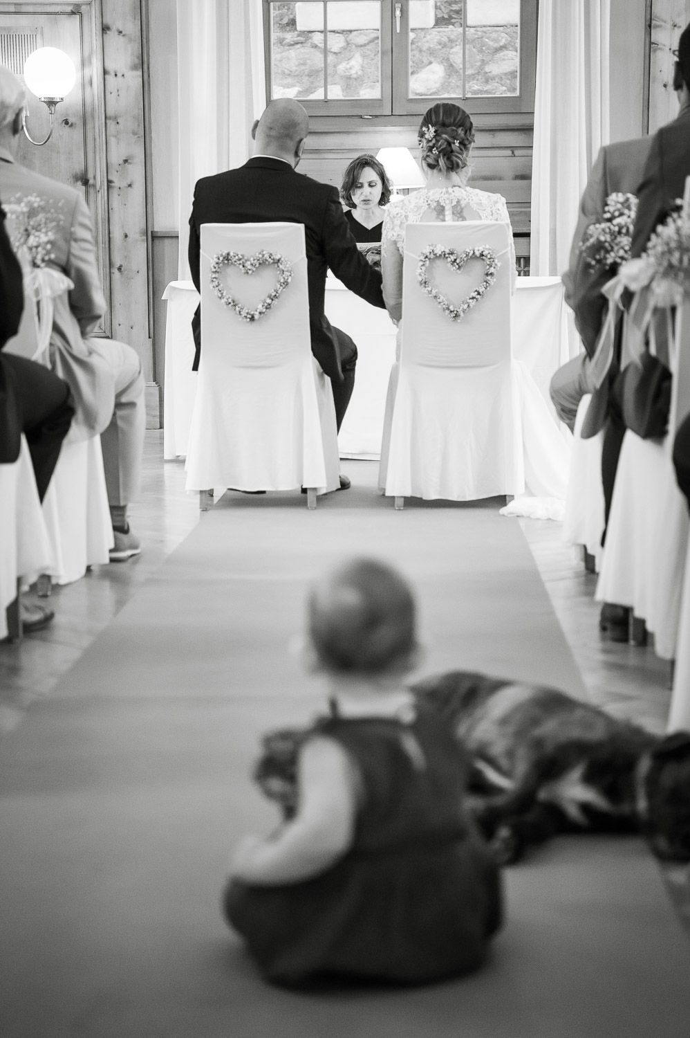 Black and white photo of wedding ceremony with couple seated at altar and baby in foreground.