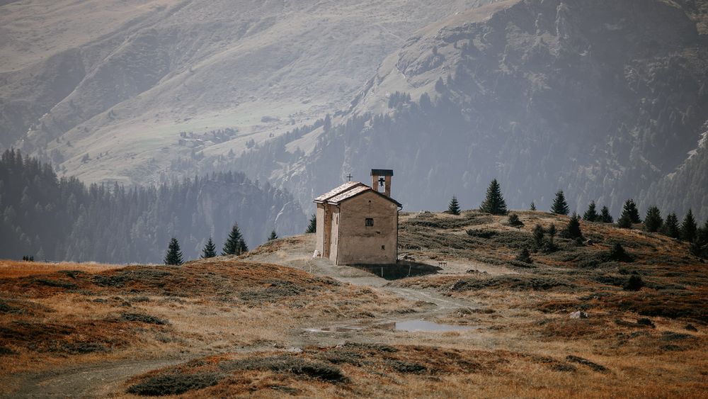 Small rustic mountain chapel on a rocky hillside surrounded by alpine trees and mountains.