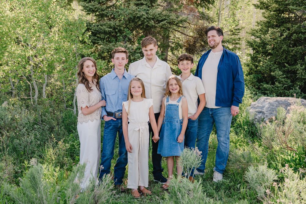 Family of seven posing outdoors in coordinating neutral outfits surrounded by lush green trees and shrubs.