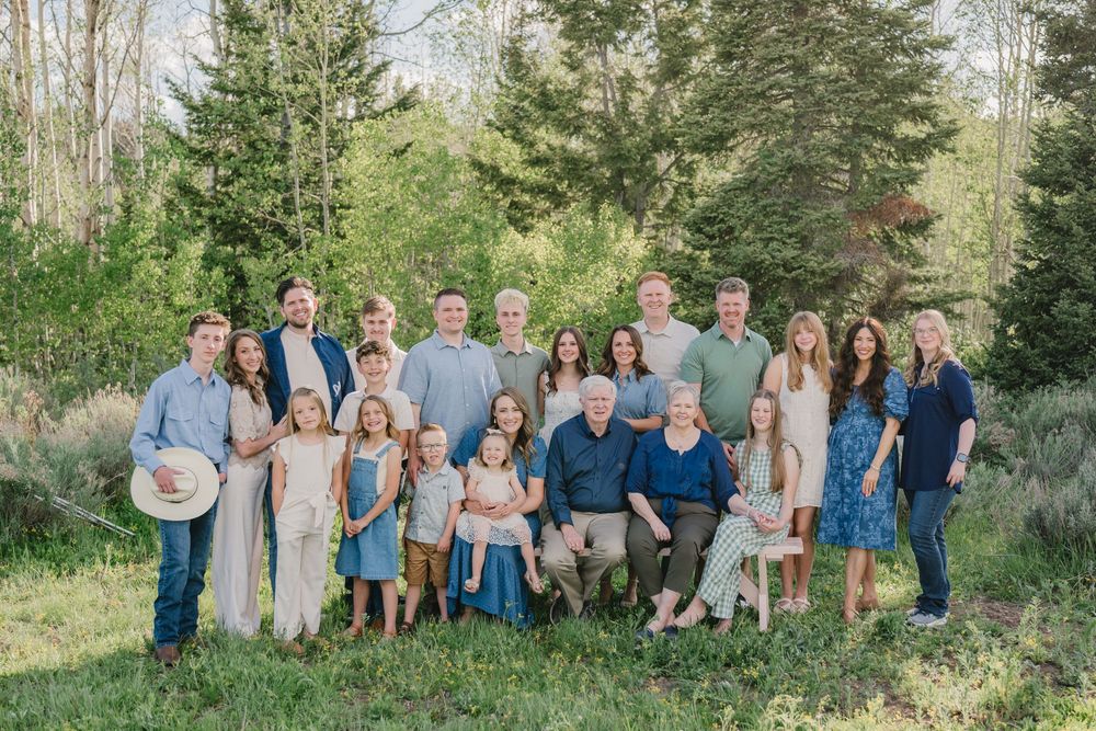 Large multigenerational family portrait outdoors in lush green setting, smiling group of adults and children in coordinated outfits.