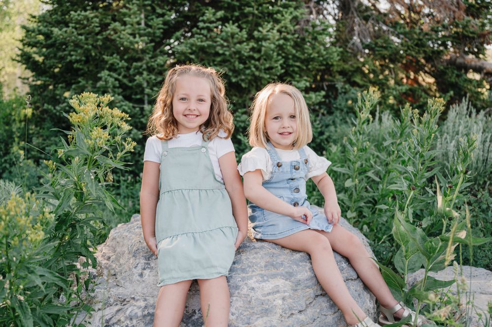 Two children sit together on a rocky outcrop with forest greenery behind them.
