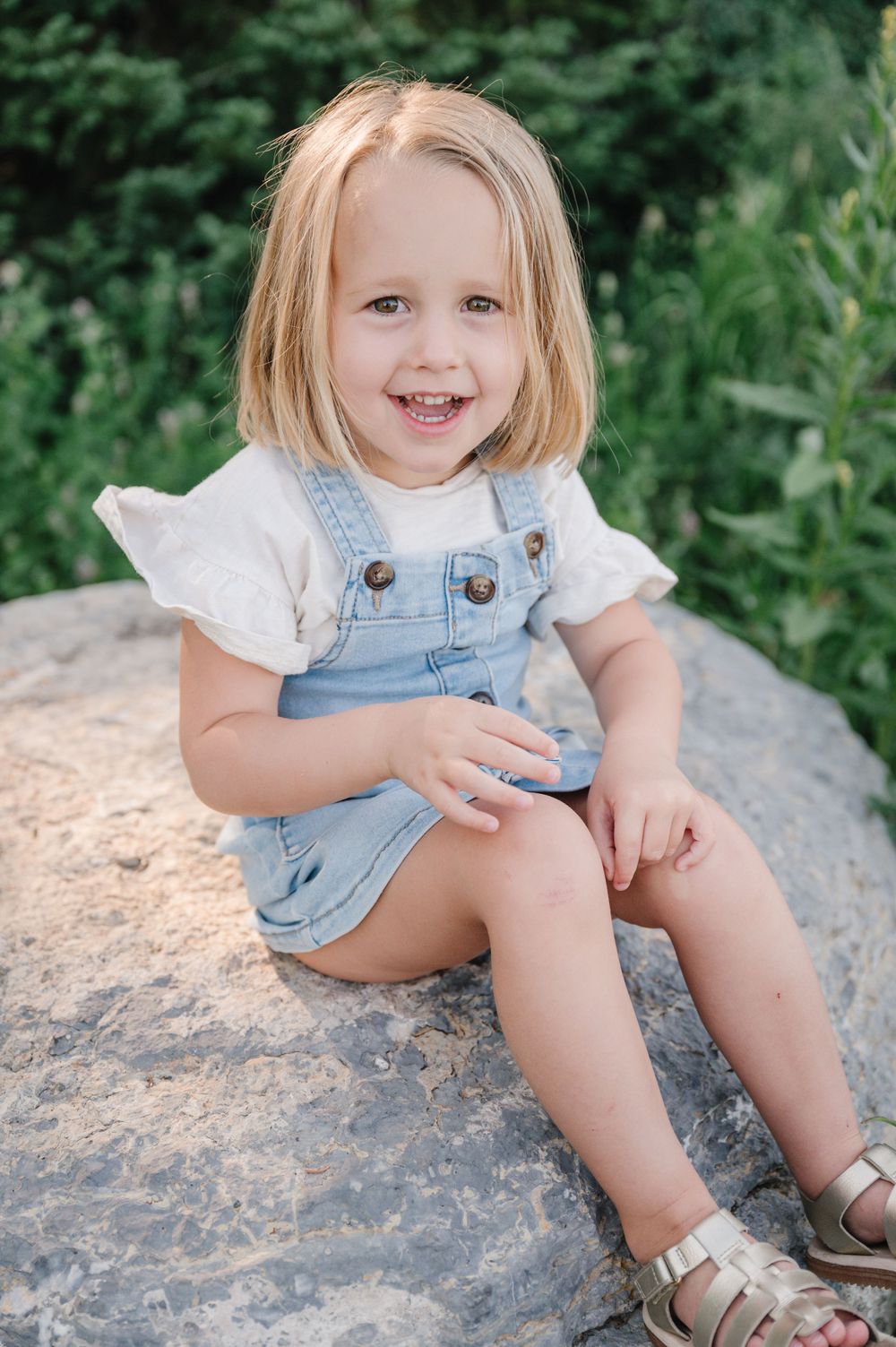 Child in denim overalls and white shirt sits on rock with bright smile.