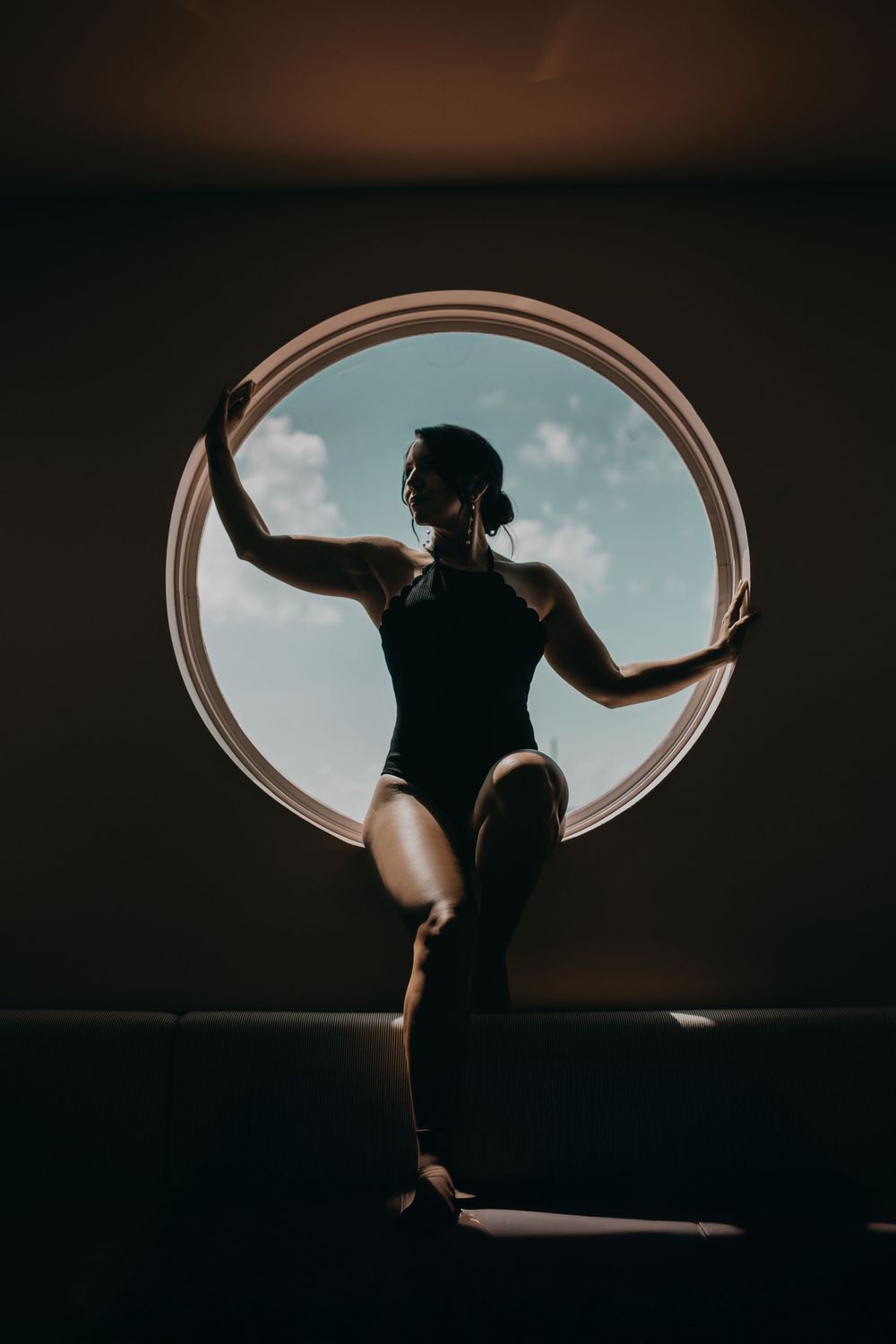 Silhouetted figure in a leotard strikes an elegant dance pose against a circular window with blue sky background.