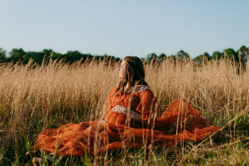 Someone in a billowing red dress sits among tall golden grass in a serene natural meadow setting.