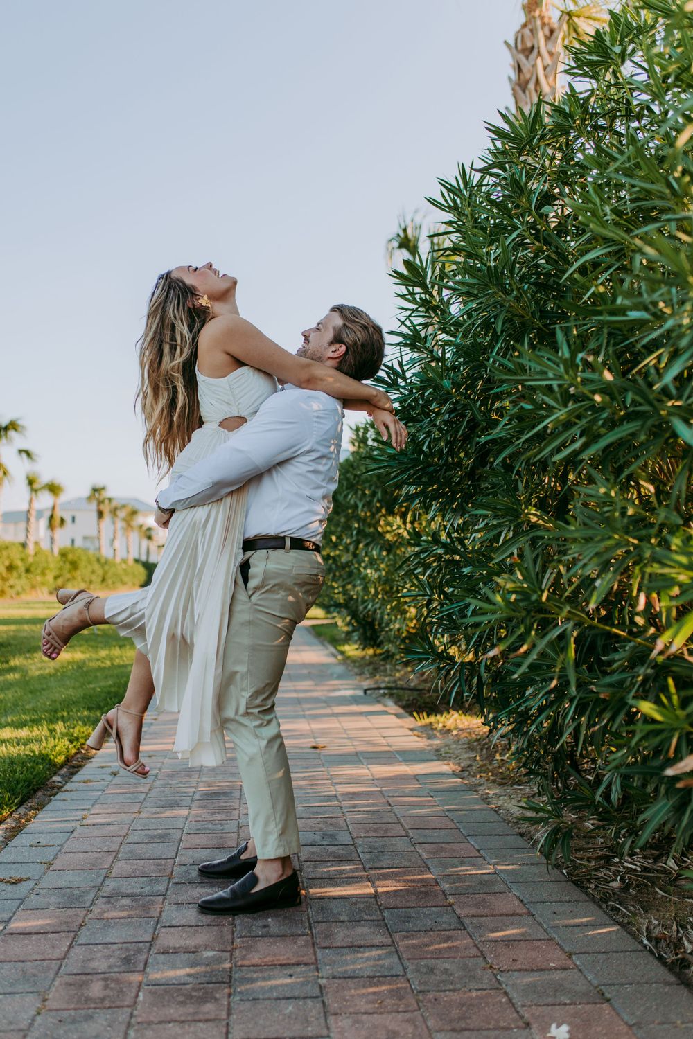 A couple embraces joyfully on a brick pathway with palm trees and lush greenery in the background.