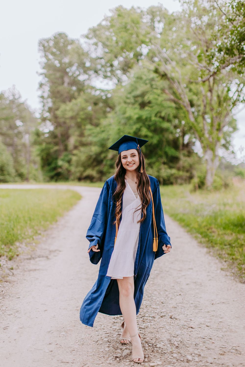 A graduate wearing a blue cap and gown poses on a dirt path surrounded by green trees and nature.