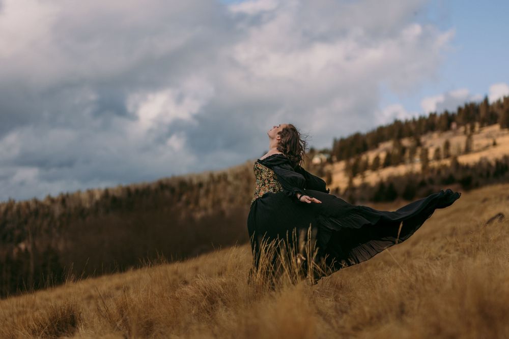 A person in a dark flowing dress stands on a windswept hillside against a cloudy sky during golden hour.