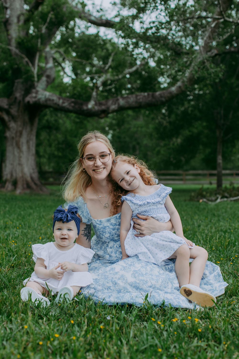Three people sitting on green grass wearing blue and white dresses on a sunny day in a park with large trees.