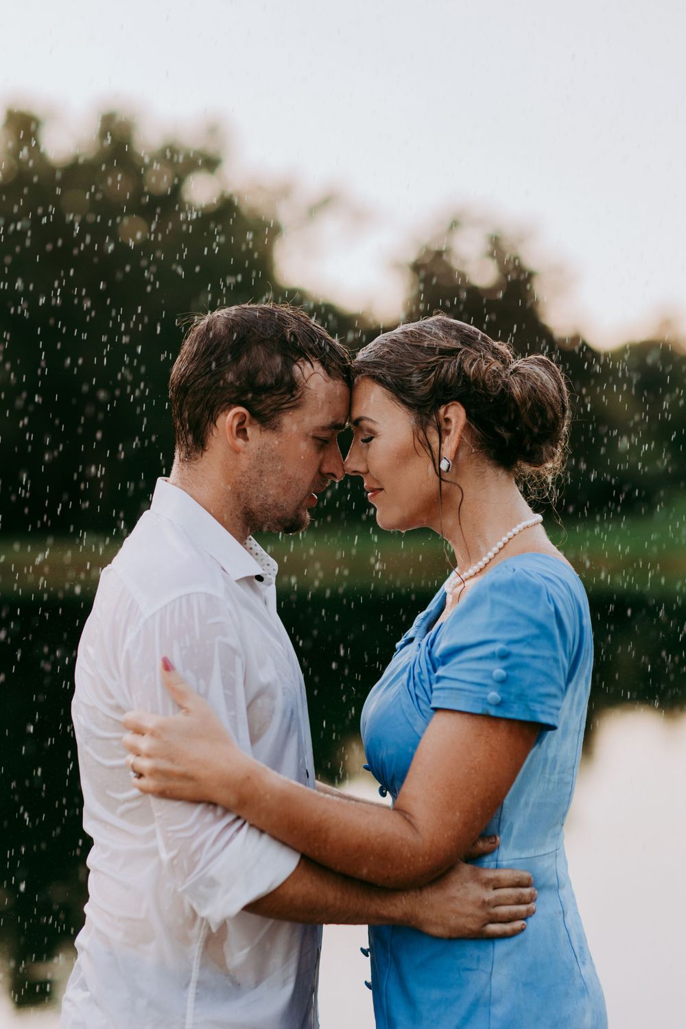 A romantic couple embraces in a light rain shower with sunlight, wearing white and blue outfits against a blurred nature backdrop.
