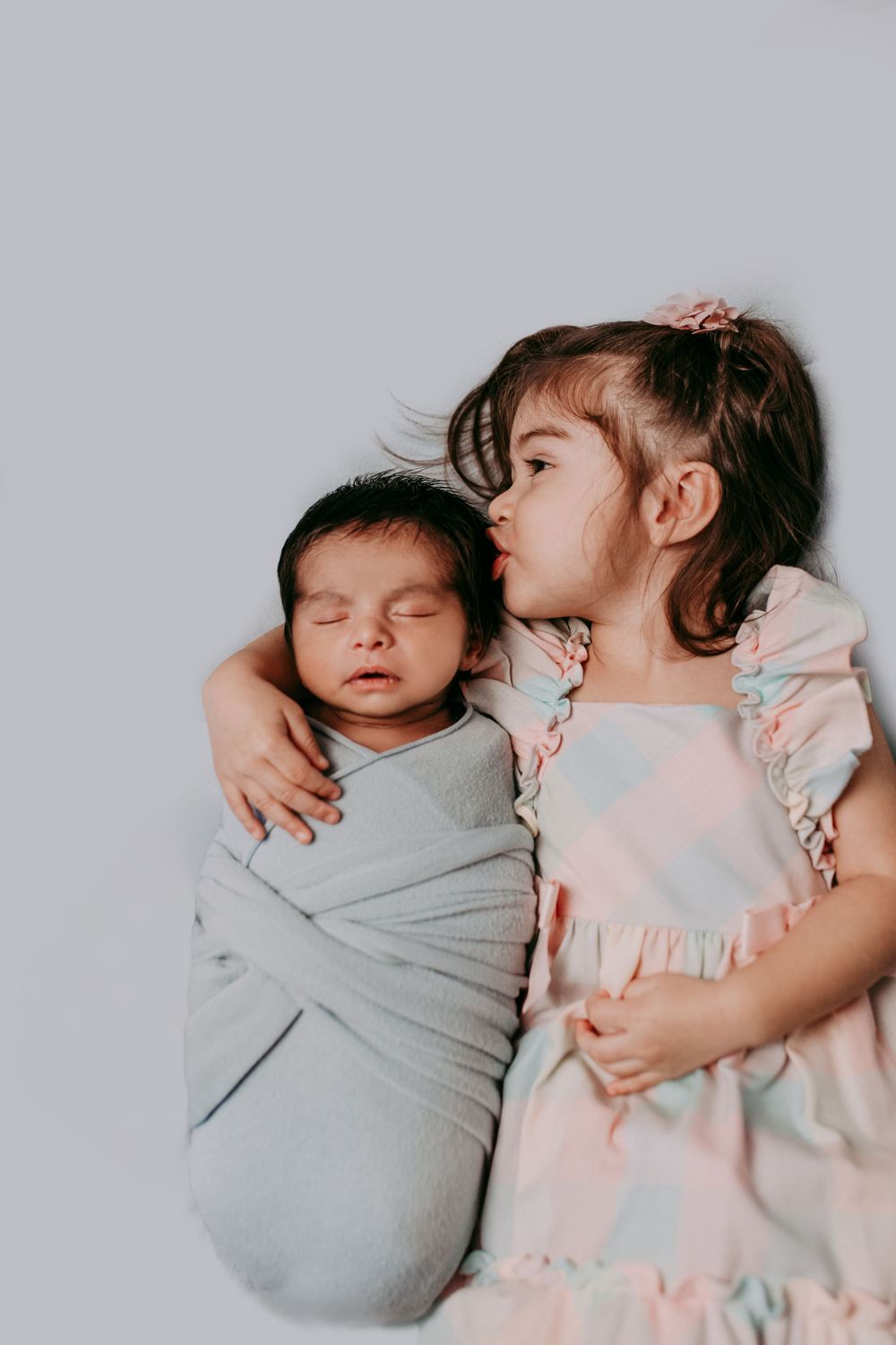 A young child in a pink dress lovingly embraces a sleeping newborn swaddled in gray.
