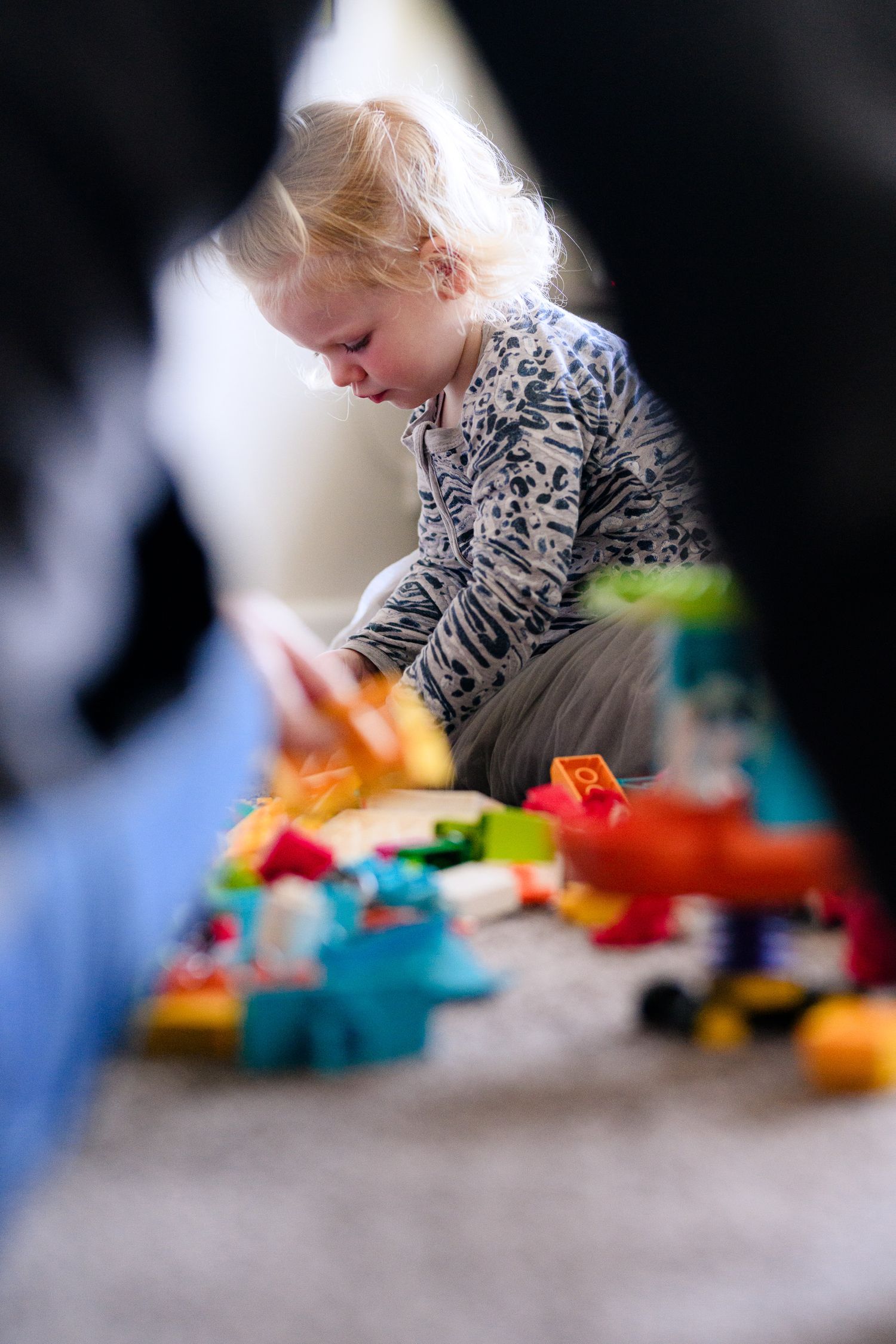 A young child in a patterned top plays with colorful toys scattered on the floor.