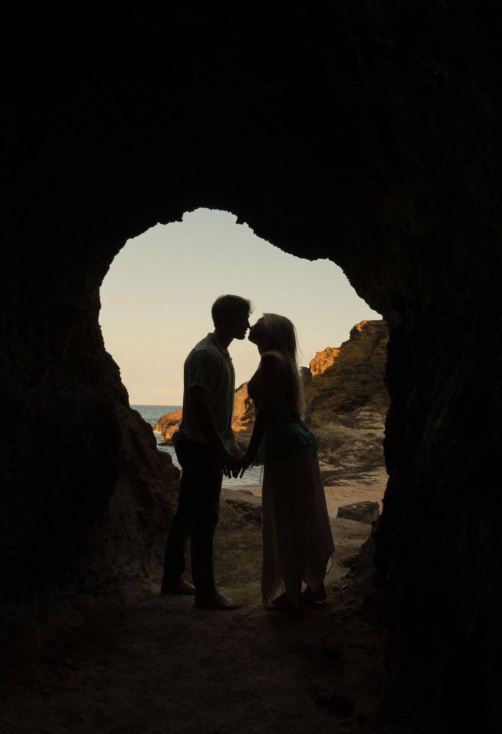 Silhouette of couple kissing inside a cave opening at sunset with ocean view in the background.
