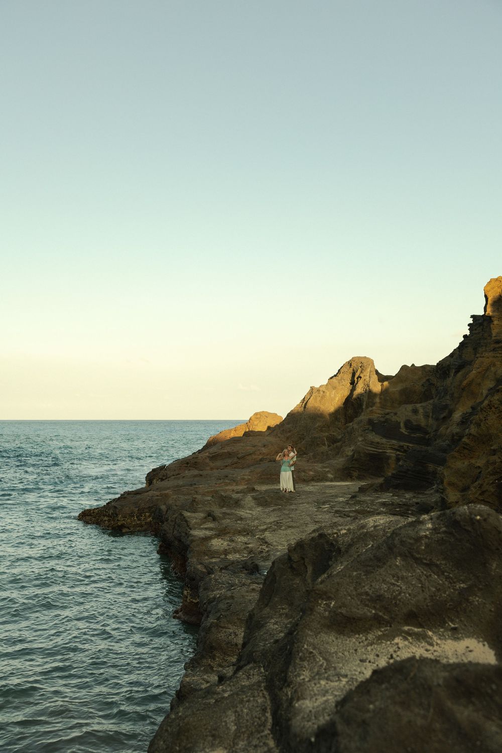A small figure stands on rugged coastal cliffs overlooking a tranquil ocean at sunset with golden light illuminating the rocks.