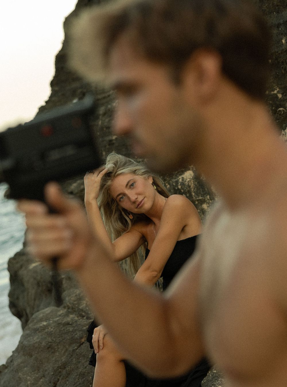 A photo shoot on rocky terrain features a person behind the camera photographing a model in black clothing at sunset.