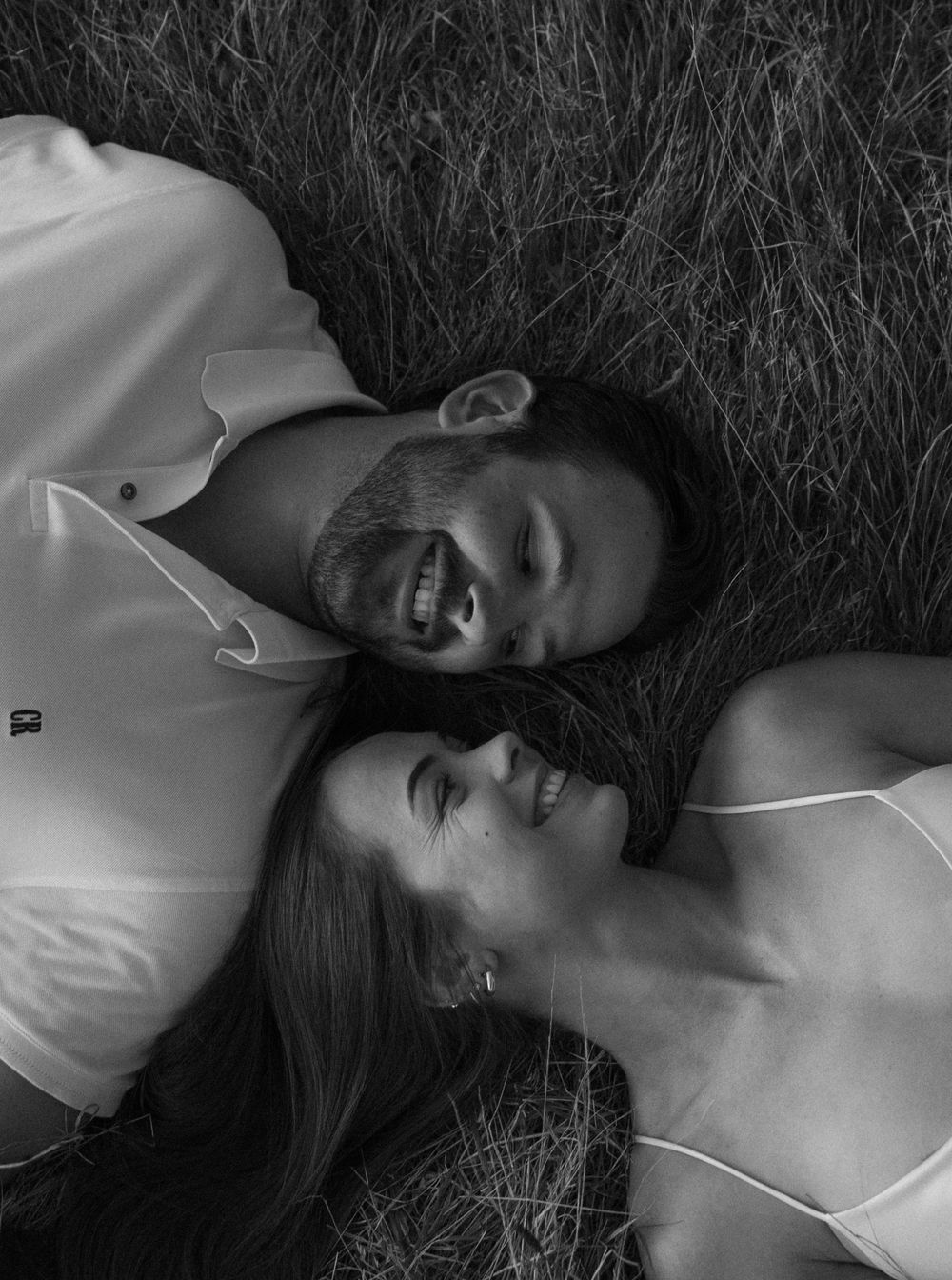 A black and white photo of a couple lying in long grass, smiling while head to head.