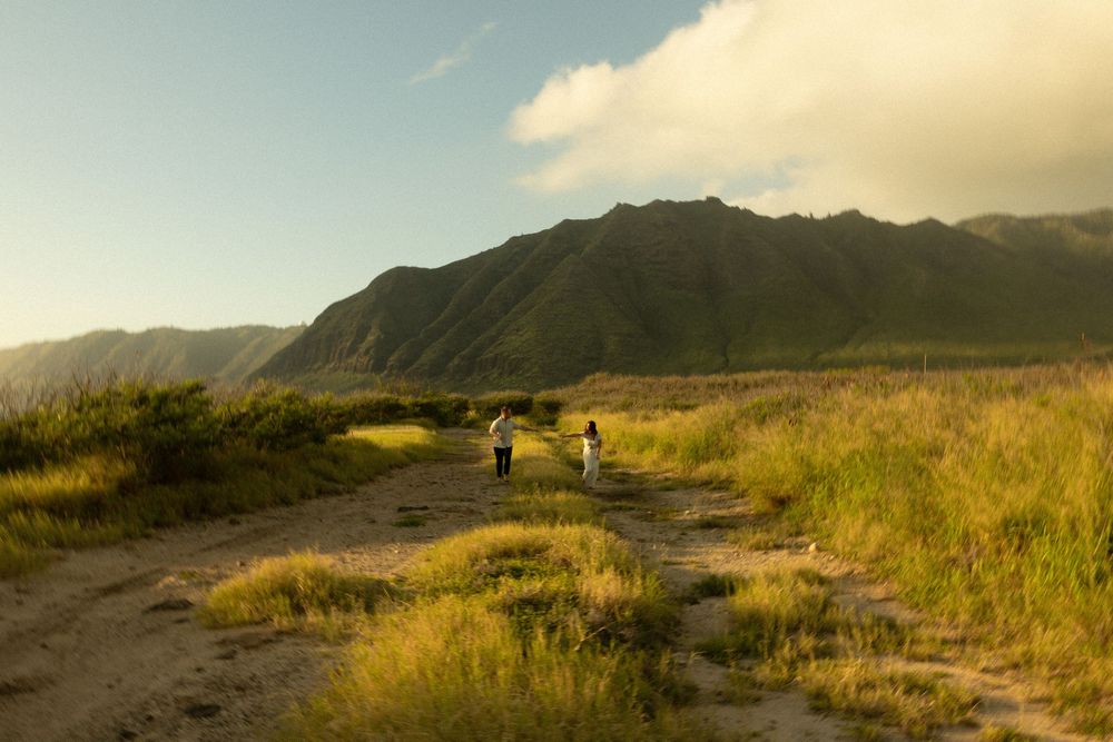 Two people hiking on a dirt trail through golden grasslands with dramatic mountain ridges in the background at sunset.