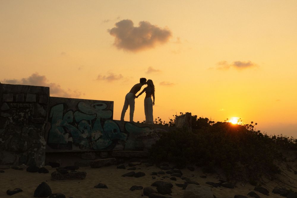 Silhouetted figures lean in for a romantic kiss against a golden sunset sky on a stone wall.