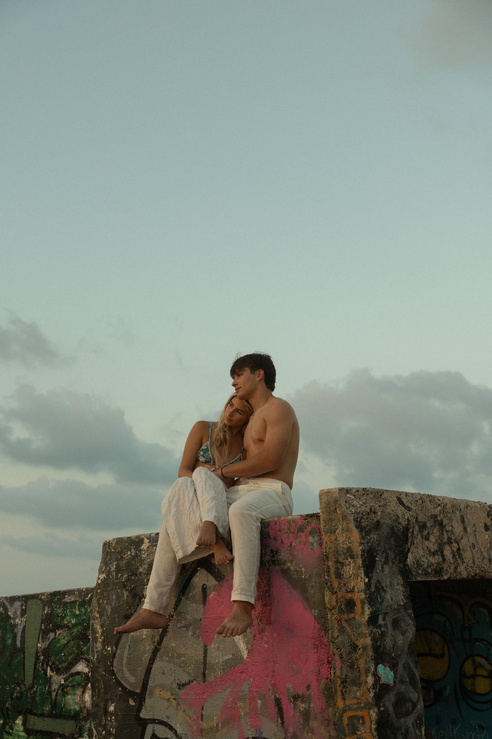 Two people share an intimate moment while sitting on a graffiti-covered concrete wall at sunset.