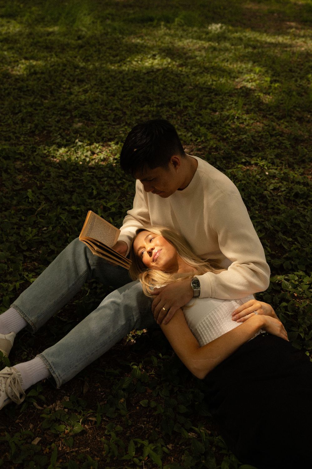 A couple relaxes on grass at sunset with one person reading while the other rests their head on their lap.