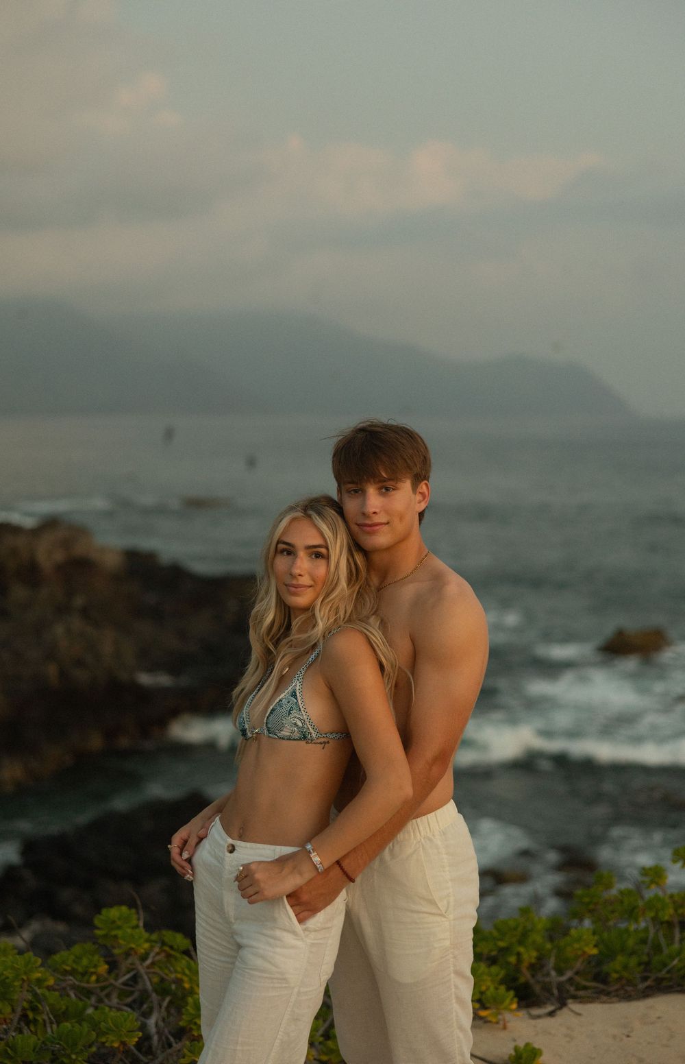 A couple embraces on a rocky Hawaiian beach during sunset with mountains and ocean waves in the background.