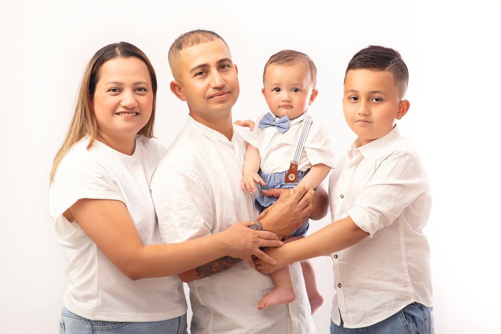 Family of four posing for portrait, parents holding baby in bow tie, older son beside them, white background.