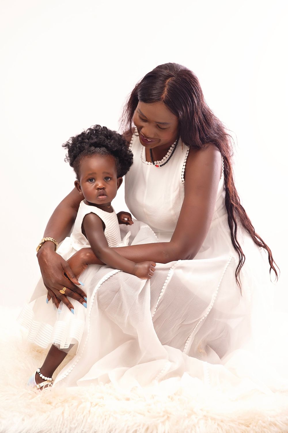 A mother and baby girl in matching white dresses sit together on a fur rug in a bright studio portrait.