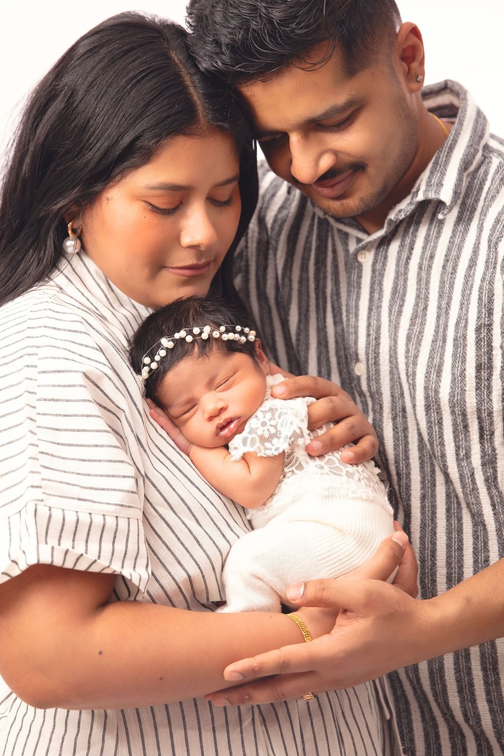 New parents tenderly cradling their sleeping newborn baby dressed in white lace, sharing a loving family portrait.