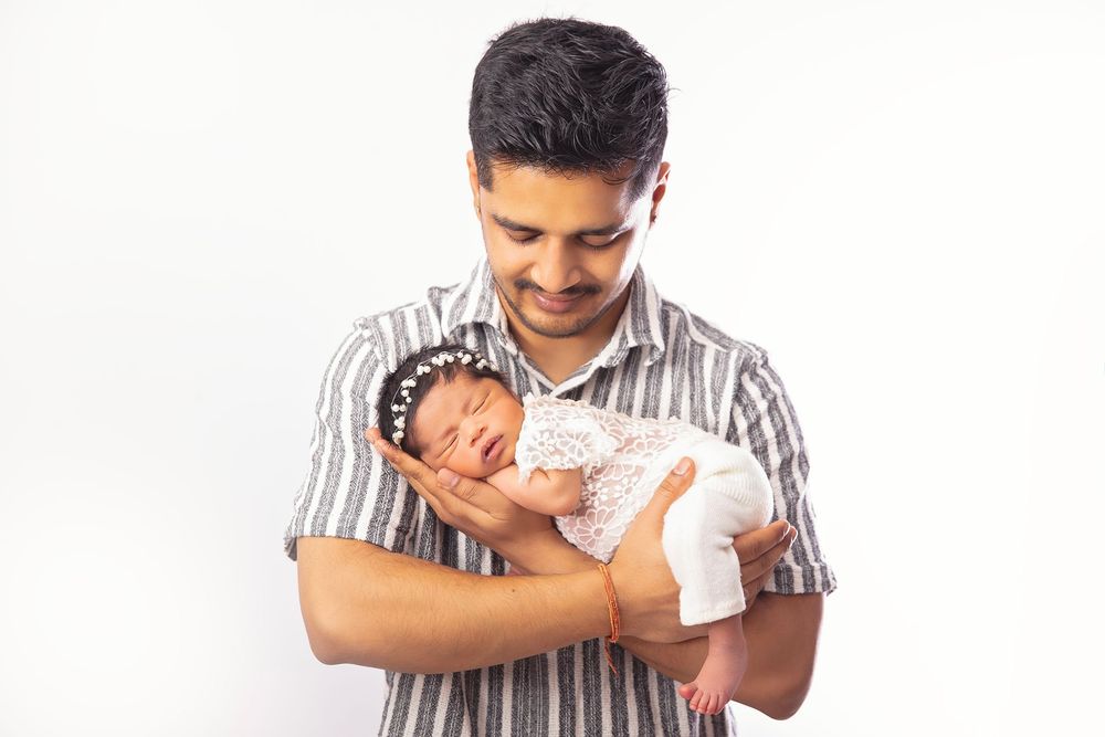 A young man in a striped shirt smiles while gently cradling a sleeping newborn baby against a white background.