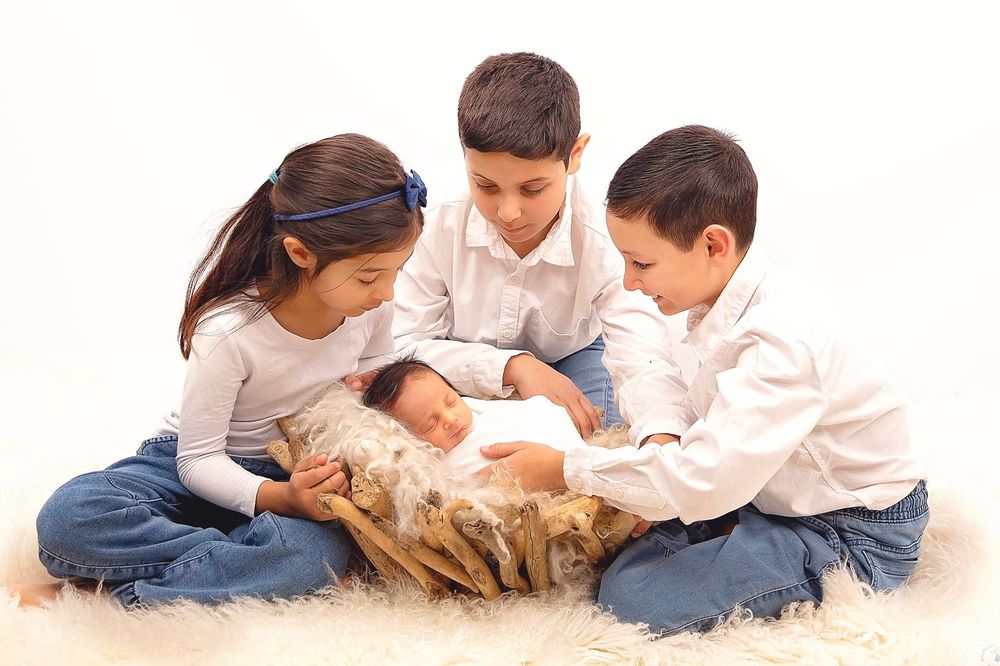 Three children sitting on a fluffy rug, lovingly gazing at a sleeping newborn baby nestled in a wooden bowl.