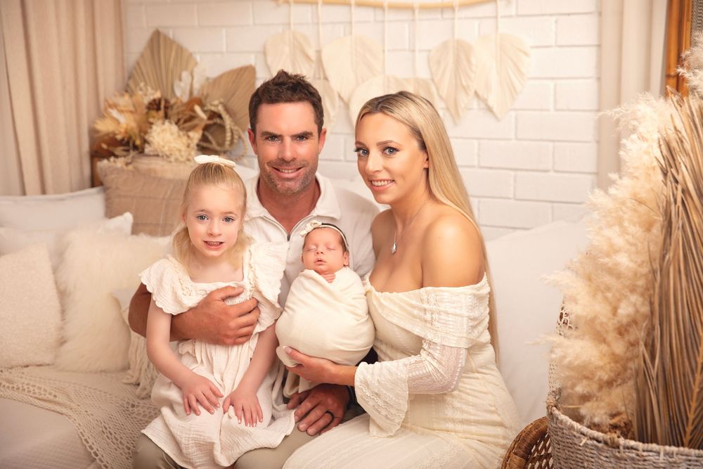 Family of four poses for a newborn photo shoot in a boho-styled studio with warm neutral tones.