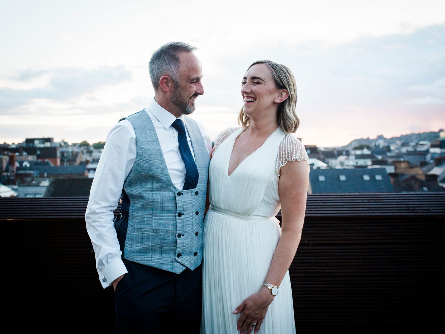 A joyful couple shares a candid moment on a rooftop during sunset, with a blurred cityscape in the background.