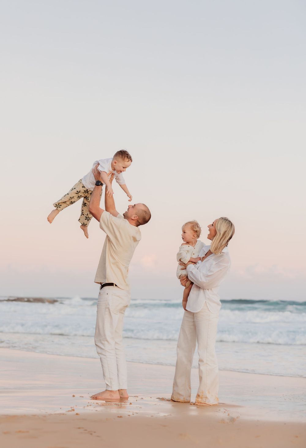 A dad holding his son up to do an airplane pose, mum holding brother and watching on during their relaxed golden hour family session.