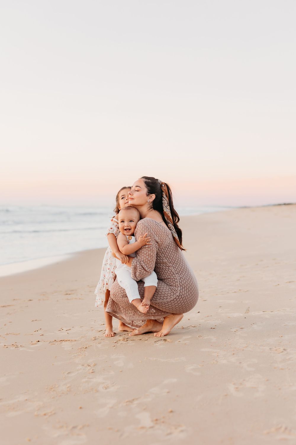 A mum gently cuddling her two kids on the beach at sunrise golden hour on the sand.