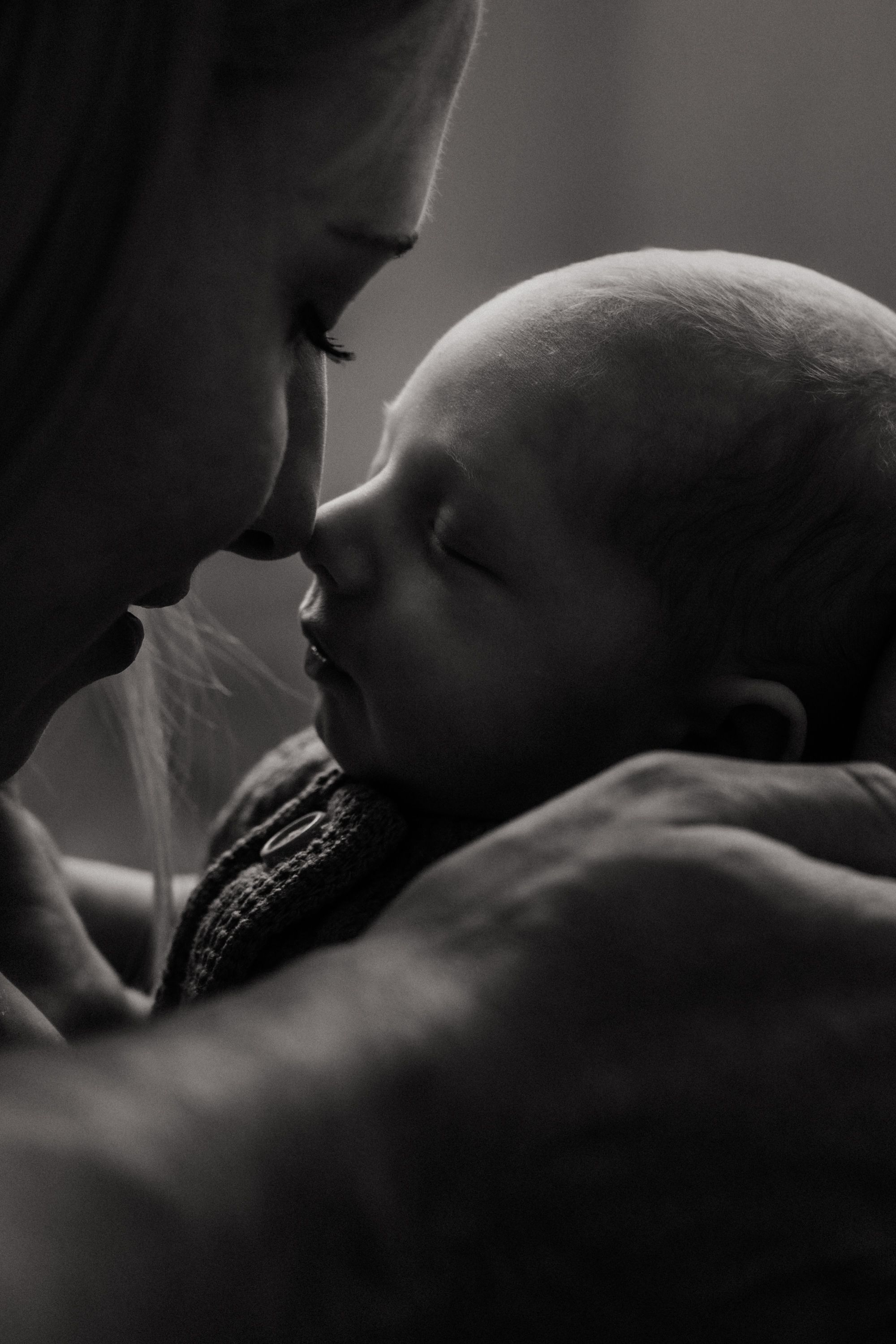 Intimate black and white portrait showing tender moment between parent and newborn baby in soft lighting.