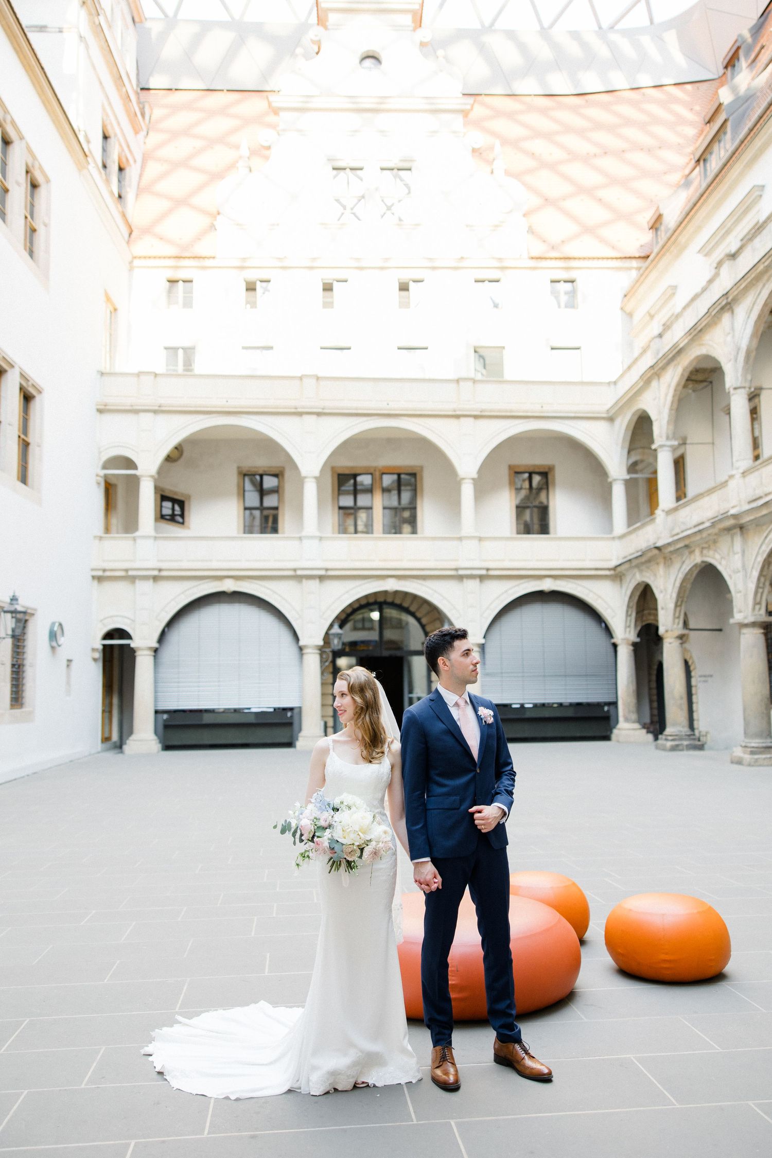 A bride and groom pose in an elegant courtyard with white arches and orange decorative spheres behind them.