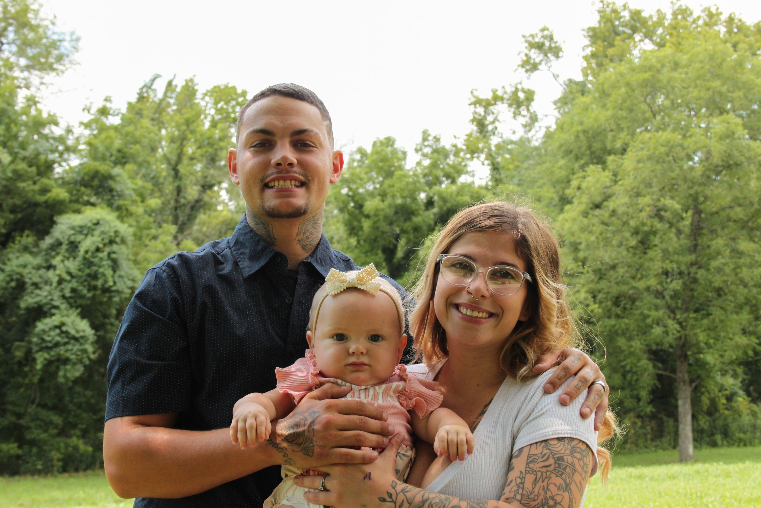 Mom and dad holding baby smiling at stockstill park in front of trees.