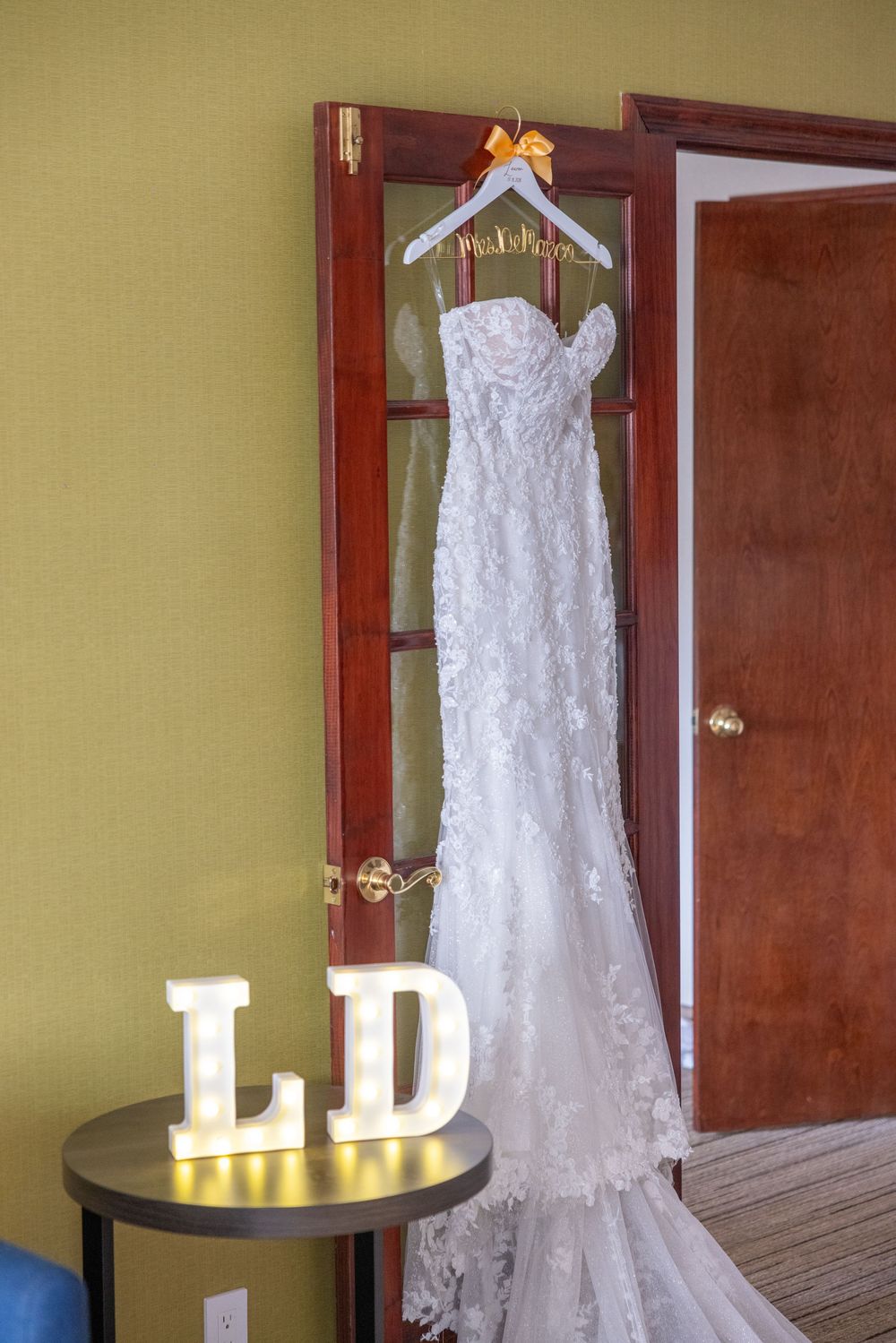 White lace wedding dress hanging on a door with illuminated LD marquee letters on a side table below.