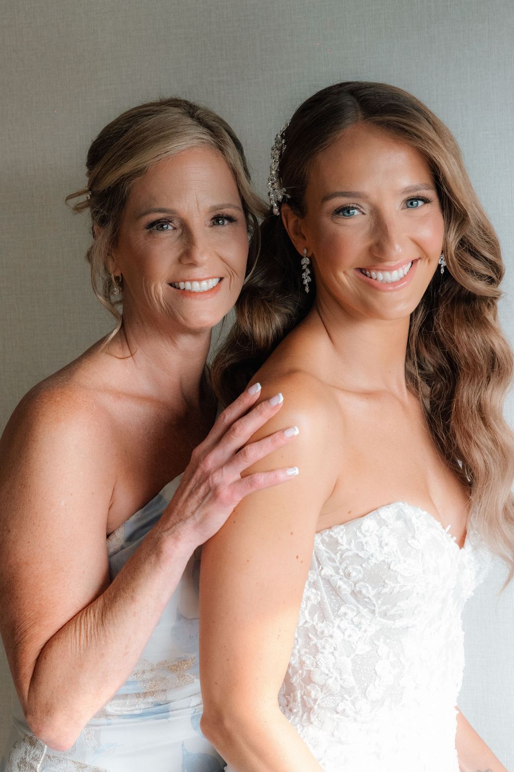 Smiling bride in strapless lace gown poses with older woman in blue dress during wedding preparations.