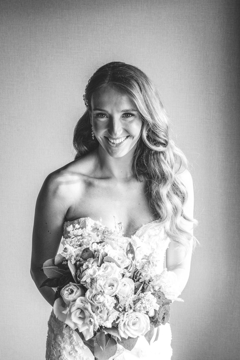 Black and white portrait of a smiling bride holding a floral bouquet in a strapless lace wedding gown.