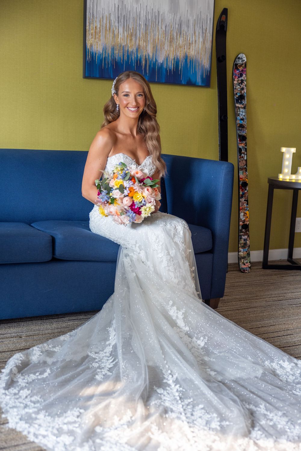 Radiant bride in strapless lace gown with long train sits on blue sofa holding colorful floral bouquet.
