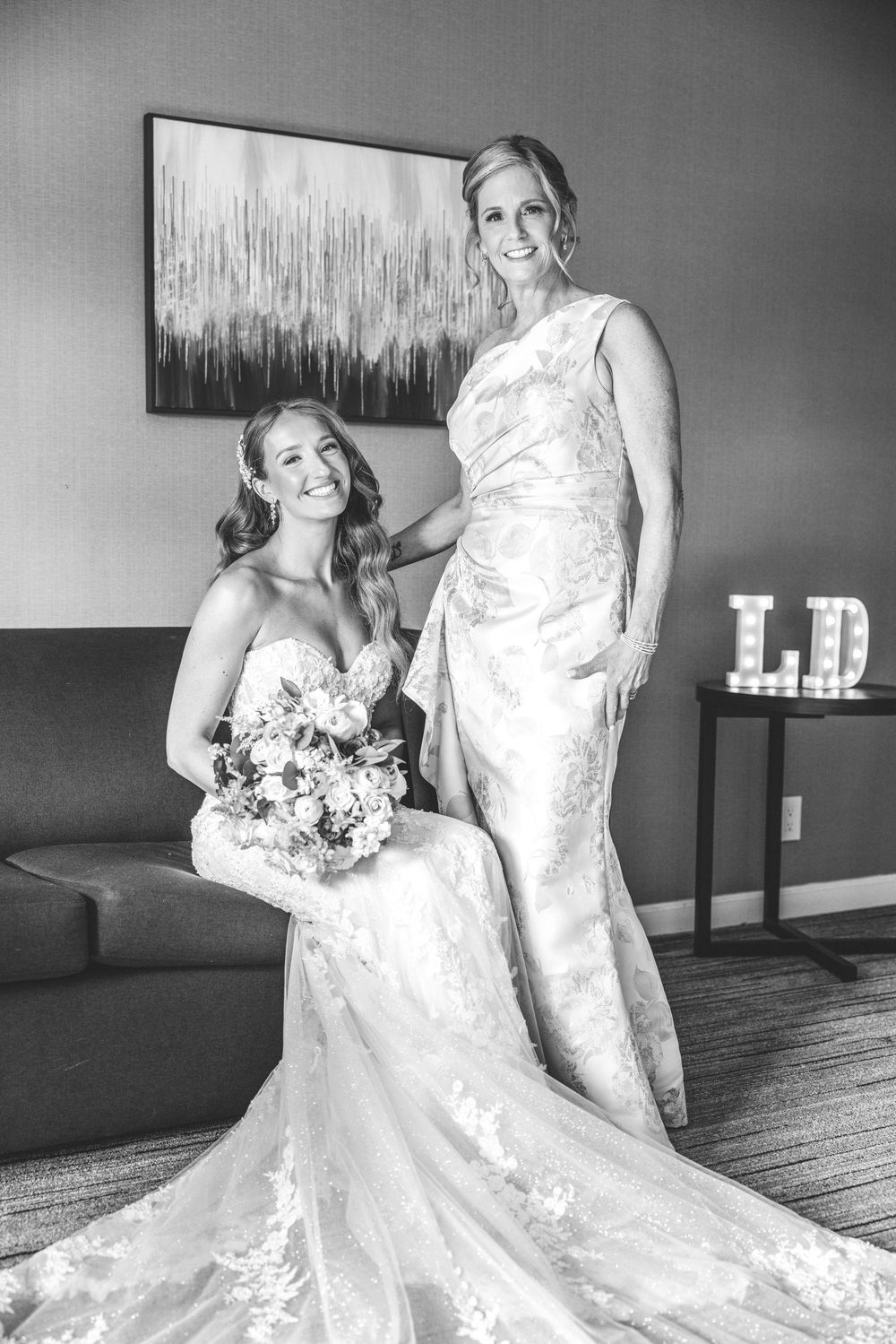 Black and white photo of bride seated with bouquet and older woman standing beside her in elegant gown.